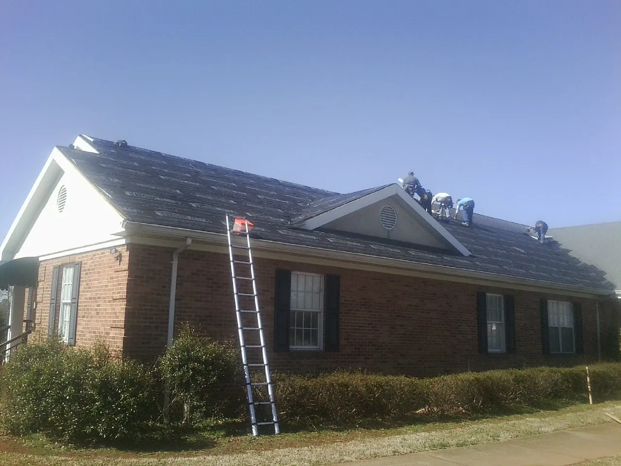 Crew tearing shingles off a brick ranch roof with a ladder set — Emergency Roof Repair work by our crew in Gilsum