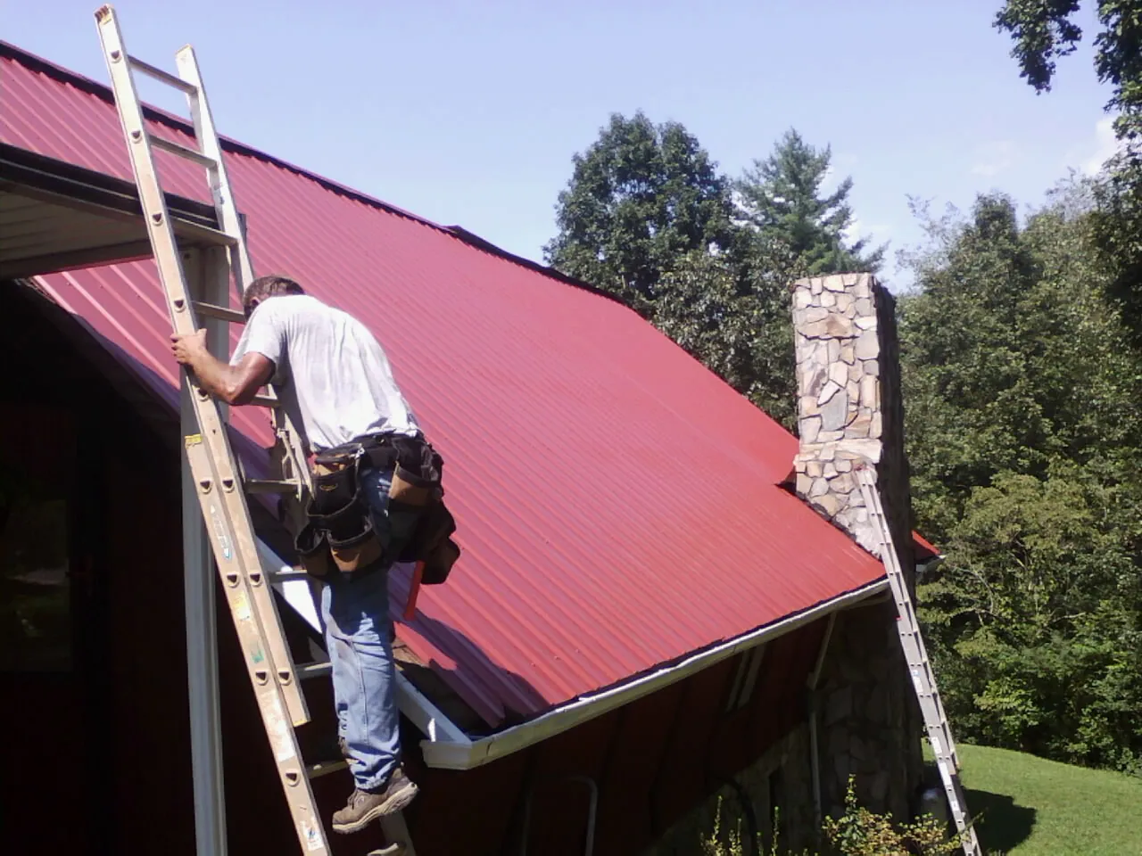Roofer on a ladder beside a red metal roof and stone chimney — Soffit & Fascia Repair by Roofing Brothers in Gilsum